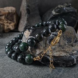 Può includere: Un braccialetto di perline verde scuro con chiusura e catena color oro. Il braccialetto è composto da grandi perline rotonde ed è esposto su una superficie di pietra grigio scuro. Lo sfondo presenta rocce ruvide grigio scuro.