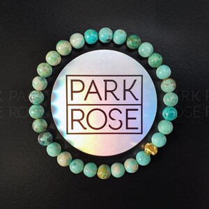 May include: A turquoise beaded bracelet with a gold accent bead. The bracelet is displayed on a black surface with a circular holographic sticker in the center that reads "PARK ROSE" in a black font.