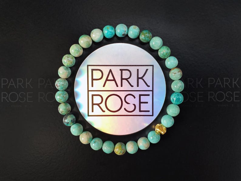May include: A turquoise beaded bracelet with a gold accent bead. The bracelet is displayed on a black surface with a circular holographic sticker in the center that reads "PARK ROSE" in a black font.