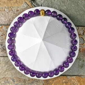 May include: A purple amethyst gemstone bracelet with a gold bead accent. The bracelet is on a white background.