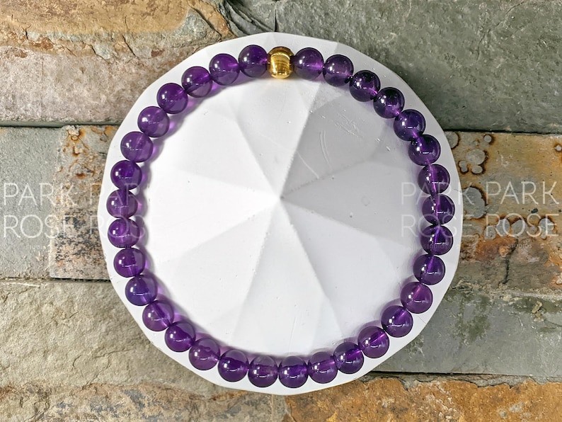 May include: A purple amethyst gemstone bracelet with a gold bead accent. The bracelet is on a white background.