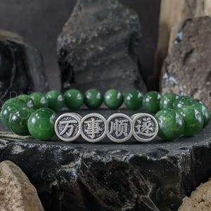 May include: A green jade bead bracelet with four silver-colored circular charms. Each charm has an engraved Chinese character. The bracelet is displayed on a dark, textured surface with dark rock formations in the background.