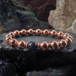 Może przedstawiać: Bransoletka z koralików miedzianych z pojedynczym czarnym koralikiem w środku. Bransoletka leży na ciemnoszarym tle.