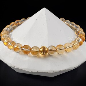 May include: A gold and amber gemstone bracelet with a gold accent bead. The bracelet is made of round beads and is displayed on a white pedestal.
