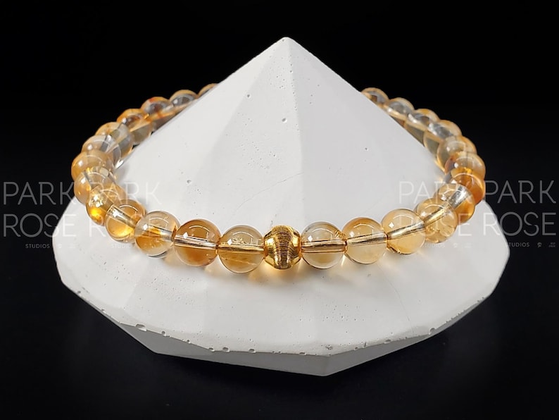 May include: A gold and amber gemstone bracelet with a gold accent bead. The bracelet is made of round beads and is displayed on a white pedestal.