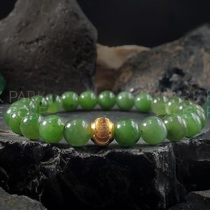 May include: A green jade bracelet with a gold bead accent. The bracelet is made of round beads and is on a dark background.