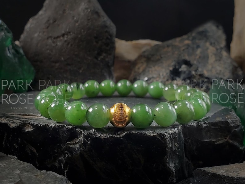 May include: A green jade bracelet with a gold bead accent. The bracelet is made of round beads and is on a dark background.