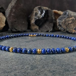 May include: A necklace featuring small, round, deep blue beads interspersed with gold-colored beads. The necklace is displayed on a dark gray surface, with dark rocks in the background. The text "PARK ROSE" is visible.