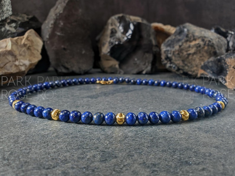 May include: A necklace featuring small, round, deep blue beads interspersed with gold-colored beads. The necklace is displayed on a dark gray surface, with dark rocks in the background. The text "PARK ROSE" is visible.