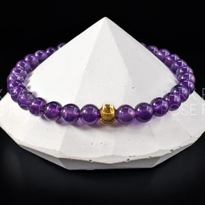 May include: A purple amethyst gemstone bracelet with a gold bead accent. The bracelet is on a white, triangular display stand.