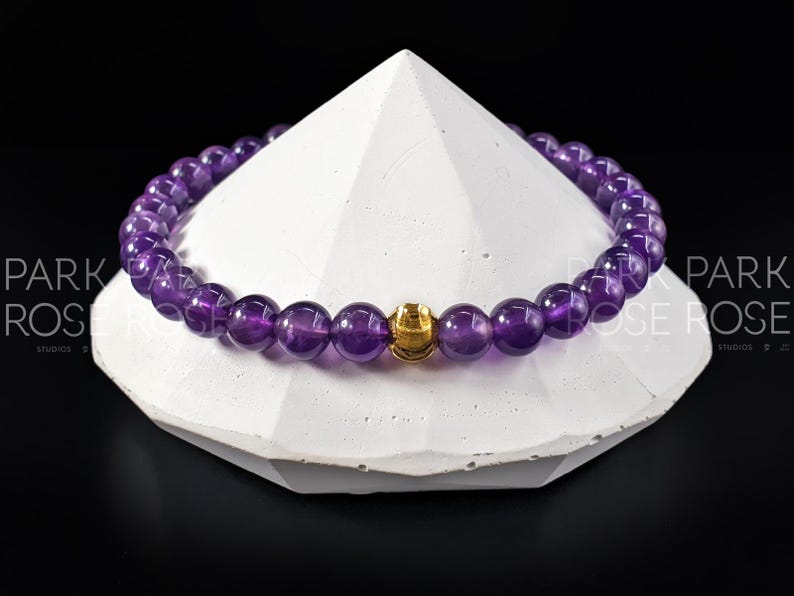 May include: A purple amethyst gemstone bracelet with a gold bead accent. The bracelet is on a white, triangular display stand.