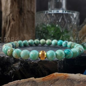 May include: A beaded bracelet featuring round, turquoise-colored beads with a gold-colored accent bead. The bracelet is displayed on a dark, textured surface, with the words "PARK PARK ROSE" visible in the background.