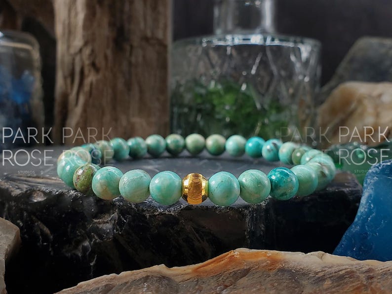 May include: A beaded bracelet featuring round, turquoise-colored beads with a gold-colored accent bead. The bracelet is displayed on a dark, textured surface, with the words "PARK PARK ROSE" visible in the background.