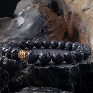 May include: A black lava rock bracelet with a gold accent bead. The bracelet is made of natural lava rock beads and has a gold-toned metal bead with a textured surface.