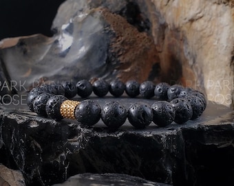 May include: A black lava rock bracelet with a gold accent bead. The bracelet is made of natural lava rock beads and has a gold-toned metal bead with a textured surface.