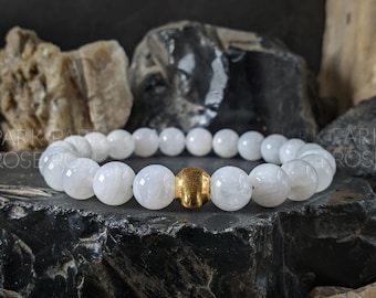 May include: White beaded bracelet with a gold-colored accent bead. The bracelet is made of round, polished white beads. The gold bead is textured. The bracelet is displayed on a dark, textured surface.