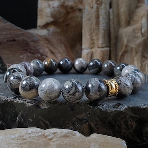 May include: A black and white patterned stone bracelet with a gold accent. The bracelet is made of round beads and is displayed on a dark gray surface.
