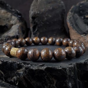 Puede incluir: Un brazalete de madera marrón oscuro con un acento dorado. El brazalete está hecho de cuentas redondas y está sobre un fondo oscuro.