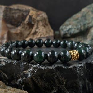 May include: A dark green beaded bracelet with a gold accent. The bracelet features large, round, polished beads. The gold accent is a cylindrical shape with a textured surface. The bracelet is displayed on a dark, textured surface.