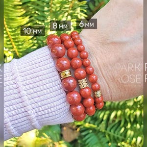 May include: Four red gemstone bracelets stacked on a wrist. The bracelets are made of round beads with gold spacers. The bracelets are labeled with their sizes: 10 mm, 8 mm, and 6 mm.