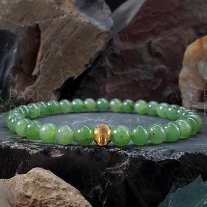 May include: A green jade bracelet with a gold bead accent. The bracelet is made of small, round beads and is shown against a dark background.