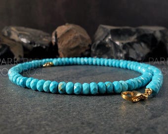 Puede incluir: Un collar de cuentas turquesas con un cierre dorado. El collar presenta cuentas redondas facetadas en un tono azul-verdoso vibrante. El cierre dorado añade un toque de elegancia. El collar se exhibe sobre una superficie gris oscuro.