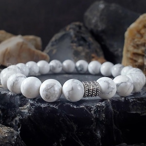May include: White howlite beaded bracelet with a silver spacer bead. The bracelet is on a dark gray background.
