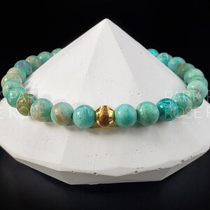 May include: A turquoise beaded bracelet with a gold-colored accent bead. The bracelet is displayed on a white geometric shape against a black background. The beads have a marbled appearance.