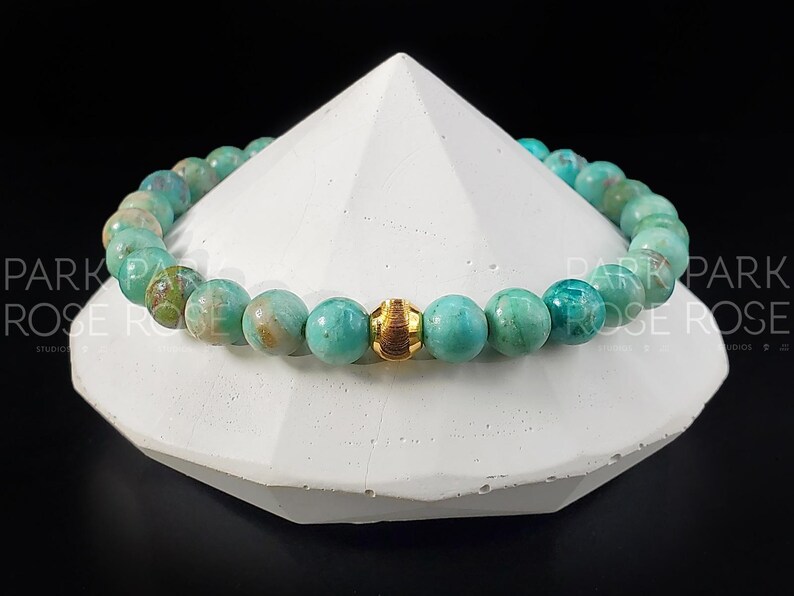 May include: A turquoise beaded bracelet with a gold-colored accent bead. The bracelet is displayed on a white geometric shape against a black background. The beads have a marbled appearance.