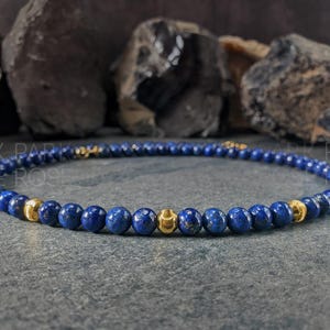 May include: A beaded necklace featuring small, round, deep blue beads interspersed with gold-colored beads. The necklace is displayed on a gray surface with dark rocks in the background. The text "PARK ROSE" is visible.