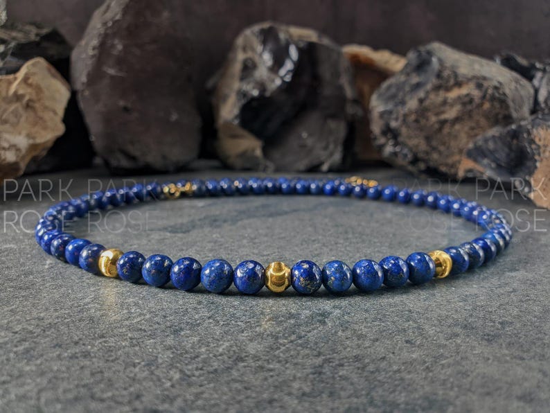 May include: A beaded necklace featuring small, round, deep blue beads interspersed with gold-colored beads. The necklace is displayed on a gray surface with dark rocks in the background. The text "PARK ROSE" is visible.