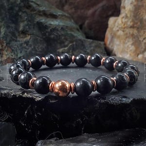 Könnte beinhalten: Schwarzes Perlenarmband mit einer Kupfer-Akzentperle. Das Armband besteht aus runden, schwarzen Perlen und einer einzelnen Kupferperle.