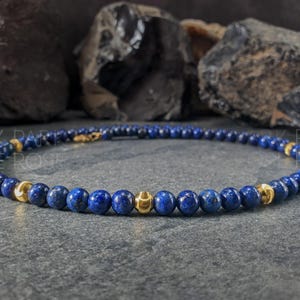 May include: A beaded bracelet featuring deep blue and gold-colored beads. The bracelet is made of small, round, blue beads interspersed with larger, gold-colored beads. The bracelet is displayed on a gray surface.
