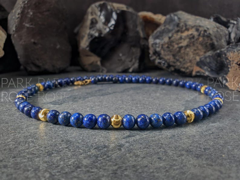 May include: A beaded bracelet featuring deep blue and gold-colored beads. The bracelet is made of small, round, blue beads interspersed with larger, gold-colored beads. The bracelet is displayed on a gray surface.