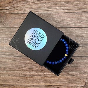 May include: A black box with the words "PARK ROSE" on a holographic sticker. The box is open, revealing a blue beaded bracelet with a gold accent. The box sits on a wooden surface.