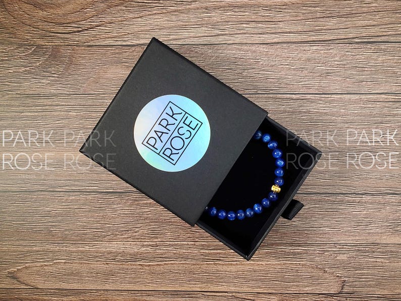 May include: A black box with the words "PARK ROSE" on a holographic sticker. The box is open, revealing a blue beaded bracelet with a gold accent. The box sits on a wooden surface.