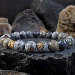 Puede incluir: Un brazalete hecho de cuentas con patrones grises y marrones con un acento dorado. El brazalete está sobre una superficie texturizada gris y marrón.