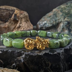 May include: A jade bead bracelet with a gold-colored Pixiu charm. The bracelet features rounded, light green beads. The Pixiu charm is a detailed, golden mythical creature. The bracelet is displayed on a dark, textured surface with rock formations in the background.