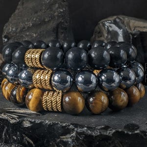 May include: Three stacked beaded bracelets. The top bracelet features large, black beads with gold-colored spacers. The middle bracelet has dark gray beads and gold spacers. The bottom bracelet has tiger's eye beads. The bracelets are displayed on a dark surface.
