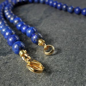 May include: Close-up of a lapis lazuli bead necklace with gold-colored clasps. The necklace features round, deep blue beads. The gold clasps are visible at the ends of the necklace, with a lobster claw clasp and a ring clasp.