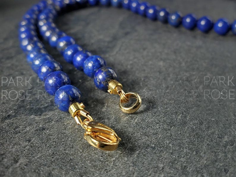 May include: Close-up of a lapis lazuli bead necklace with gold-colored clasps. The necklace features round, deep blue beads. The gold clasps are visible at the ends of the necklace, with a lobster claw clasp and a ring clasp.
