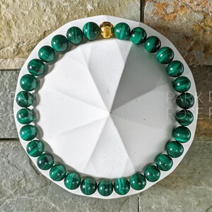 May include: A green malachite gemstone bracelet with a gold clasp. The bracelet is on a white background.