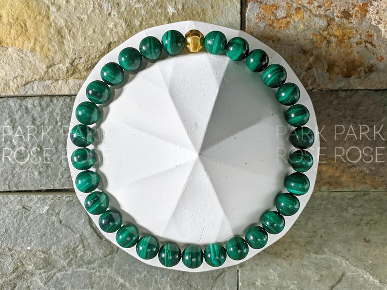 May include: A green malachite gemstone bracelet with a gold clasp. The bracelet is on a white background.