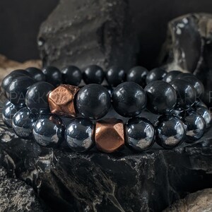 May include: Two beaded bracelets featuring black, round beads and copper-coloured geometric beads. The bracelets are stacked on a dark, textured surface, creating a contrast of colours and textures. The bracelets are a type of jewellery.