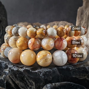 May include: Three stacked beaded bracelets with varying sizes of round, polished beads. The beads are in shades of cream, tan, brown, and red. The sizes are labeled as 6 mm, 8 mm, and 10 mm. The bracelets are displayed on a dark, textured surface.