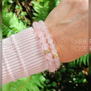 May include: Two stacked bracelets made of pink rose quartz beads with gold accents. The bracelets are worn on a person's wrist.