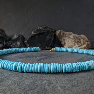 May include: A necklace composed of numerous small, flat, circular turquoise beads in a vibrant sky blue hue. The beads are strung together in a continuous strand, featuring a small gold-toned metal clasp at the center back. The text PARK ROSE is visible.