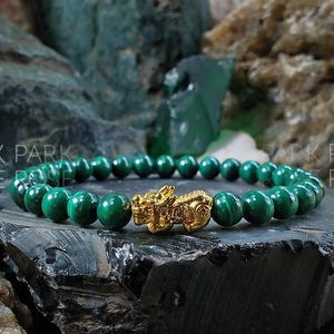May include: A green malachite beaded bracelet with a gold-toned dragon charm. The bracelet is on a dark grey surface.