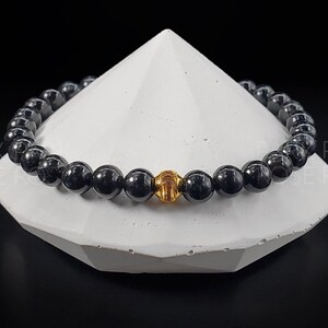 May include: A black beaded bracelet with a gold accent bead. The bracelet is displayed on a white, triangular stand.