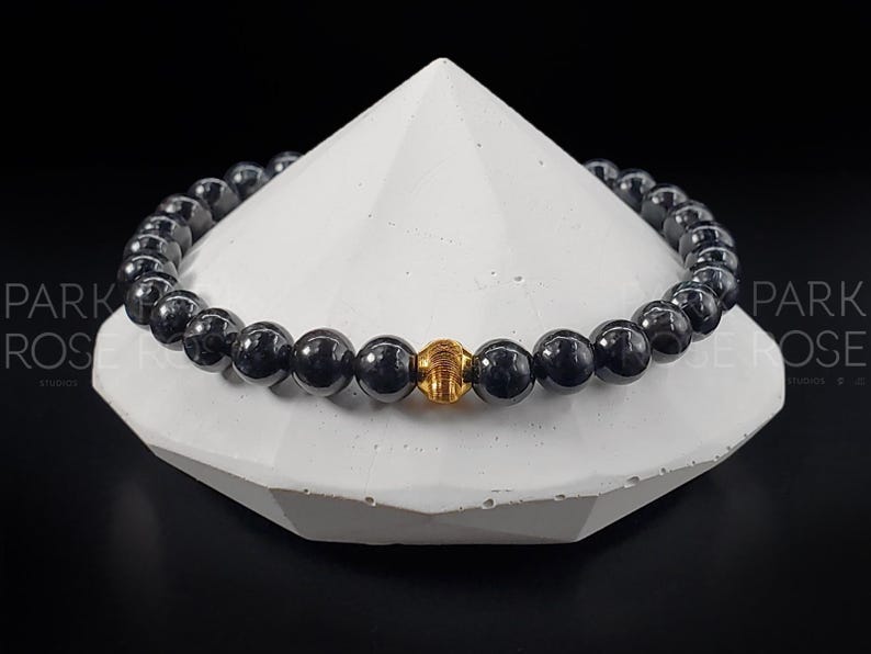May include: A black beaded bracelet with a gold accent bead. The bracelet is displayed on a white, triangular stand.